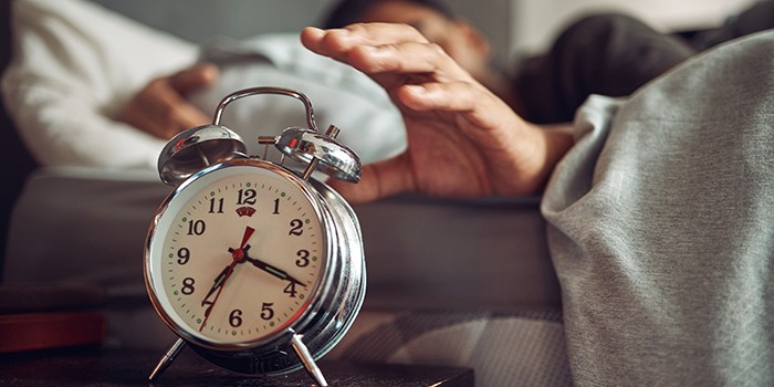 A hand reaches out from under the bedclothes to turn off the alarm clock